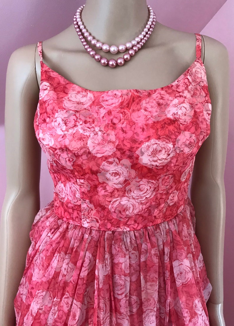 K&ouml;nnte beinhalten: Ein pinkfarbenes, &auml;rmelloses Kleid mit einem Blumendruck aus wei&szlig;en Rosen. Das Kleid hat ein tailliertes Oberteil und einen weiten Rock.