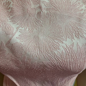 May include: Close-up of a pink fabric with a floral pattern. The fabric has a subtle shimmer and a textured surface.