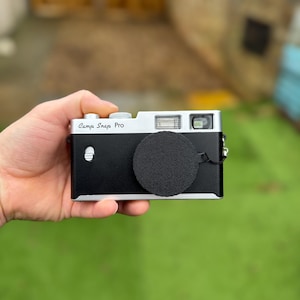 Pode incluir: Uma c&acirc;mera preta e prateada com o texto "Camp Snap Pro" &eacute; segurada em uma m&atilde;o. A c&acirc;mera tem uma tampa de lente e um flash. O fundo &eacute; grama verde desfocada e uma cerca de madeira.