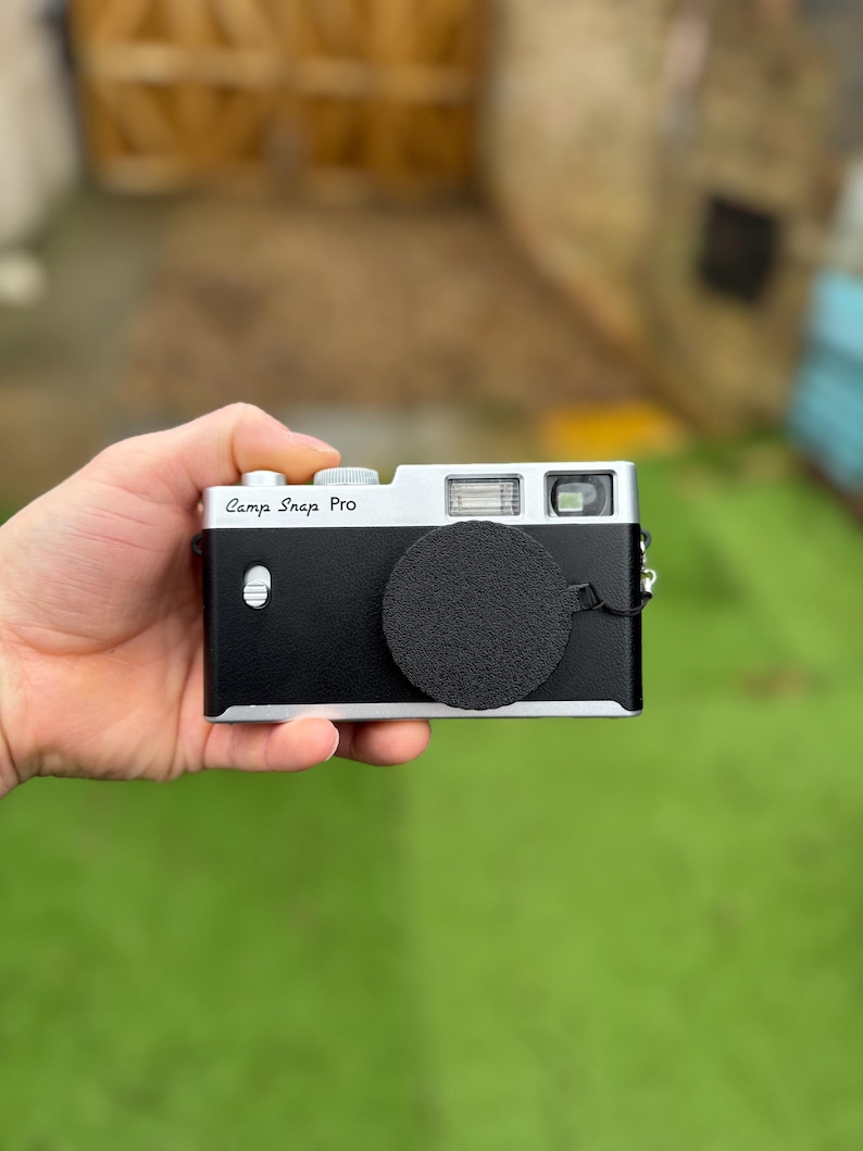 Pode incluir: Uma c&acirc;mera preta e prateada com o texto "Camp Snap Pro" &eacute; segurada em uma m&atilde;o. A c&acirc;mera tem uma tampa de lente e um flash. O fundo &eacute; grama verde desfocada e uma cerca de madeira.