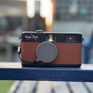 May include: A brown and black camera with a leather exterior and a grey lens cap. The camera has the text "Camp Snap" printed on the front.