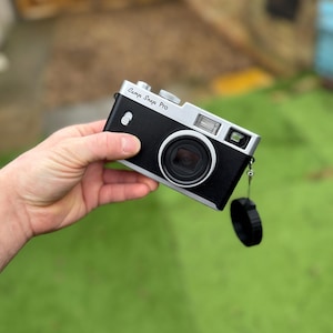 Pode incluir: Uma c&acirc;mara Camp Snap Pro preta e prateada com lente e flash. A c&acirc;mara tem uma correia de pulso e uma tampa de lente pretas. O texto "Camp Snap Pro" est&aacute; impresso na parte superior.