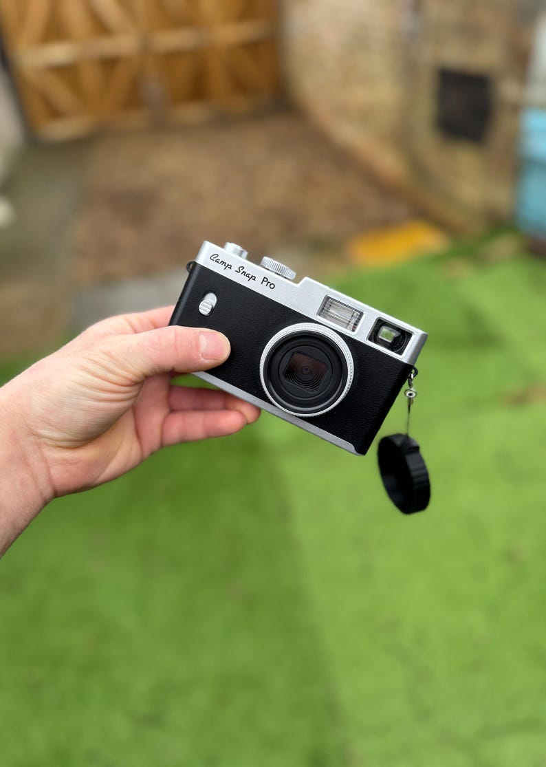 Pode incluir: Uma c&acirc;mara Camp Snap Pro preta e prateada com lente e flash. A c&acirc;mara tem uma correia de pulso e uma tampa de lente pretas. O texto "Camp Snap Pro" est&aacute; impresso na parte superior.