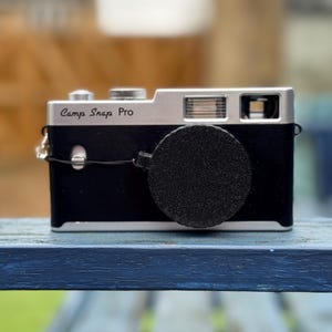 Pode incluir: C&acirc;mera Camp Snap Pro preta e prateada com tampa da lente. A c&acirc;mera est&aacute; em uma superf&iacute;cie de madeira azul. Possui flash e lente. O texto "Camp Snap Pro" &eacute; vis&iacute;vel.