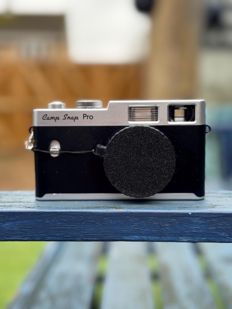 Pode incluir: C&acirc;mera Camp Snap Pro preta e prateada com tampa da lente. A c&acirc;mera est&aacute; em uma superf&iacute;cie de madeira azul. Possui flash e lente. O texto "Camp Snap Pro" &eacute; vis&iacute;vel.