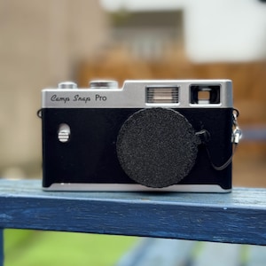 May include: A black and silver Camp Snap Pro camera with a lens cap. The camera has a silver top and a black body. The text "Camp Snap Pro" is printed on the top of the camera.