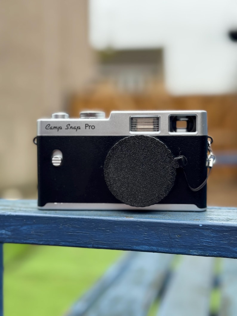 Pode incluir: Uma c&acirc;mara Camp Snap Pro preta e prateada com uma tampa de lente. A c&acirc;mara tem uma parte superior prateada e um corpo preto. O texto "Camp Snap Pro" est&aacute; impresso na parte superior da c&acirc;mara.