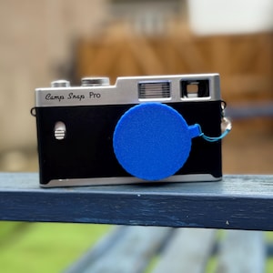 Pode incluir: Uma c&acirc;mera preta e prateada com o texto "Camp Snap Pro" na frente. Uma tampa de lente azul est&aacute; presa &agrave; c&acirc;mera. A c&acirc;mera est&aacute; em uma superf&iacute;cie azul.