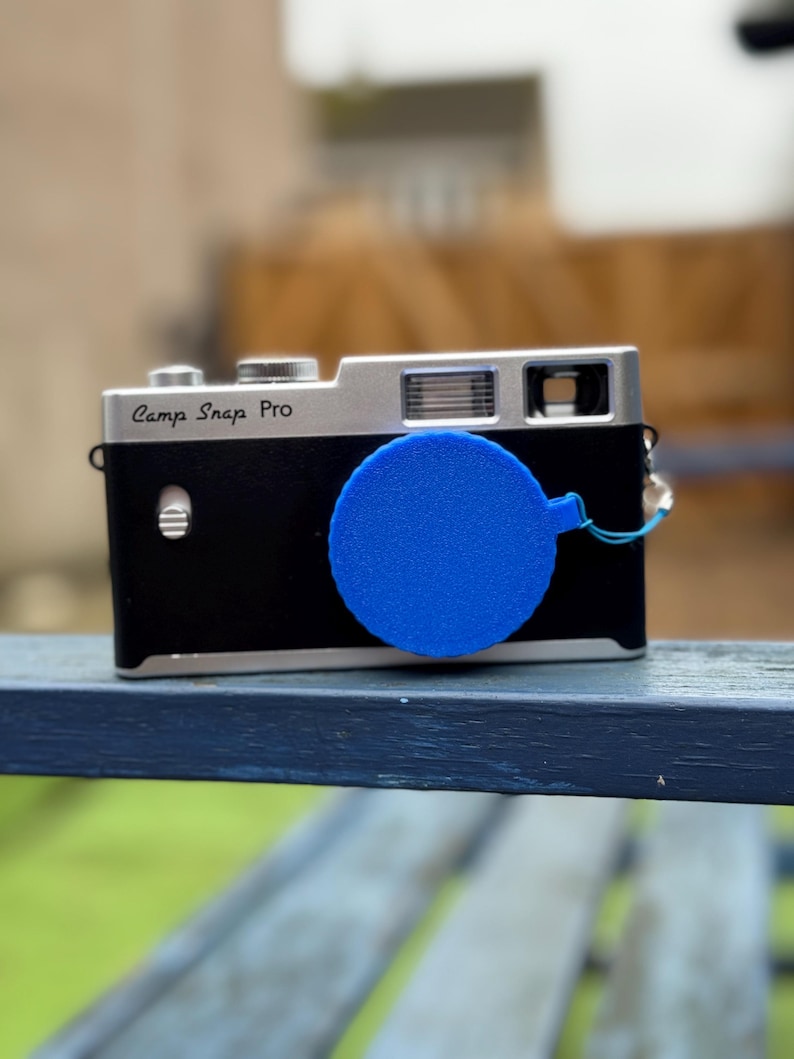 Pode incluir: Uma c&acirc;mera preta e prateada com o texto "Camp Snap Pro" na frente. Uma tampa de lente azul est&aacute; presa &agrave; c&acirc;mera. A c&acirc;mera est&aacute; em uma superf&iacute;cie azul.