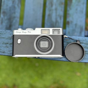 Pode incluir: Uma c&acirc;mera Camp Snap Pro preta e prateada com uma tampa de lente. A c&acirc;mera tem uma parte superior prateada e uma lente proeminente. O texto "Camp Snap Pro" est&aacute; impresso na frente. A c&acirc;mera est&aacute; em uma superf&iacute;cie azul.