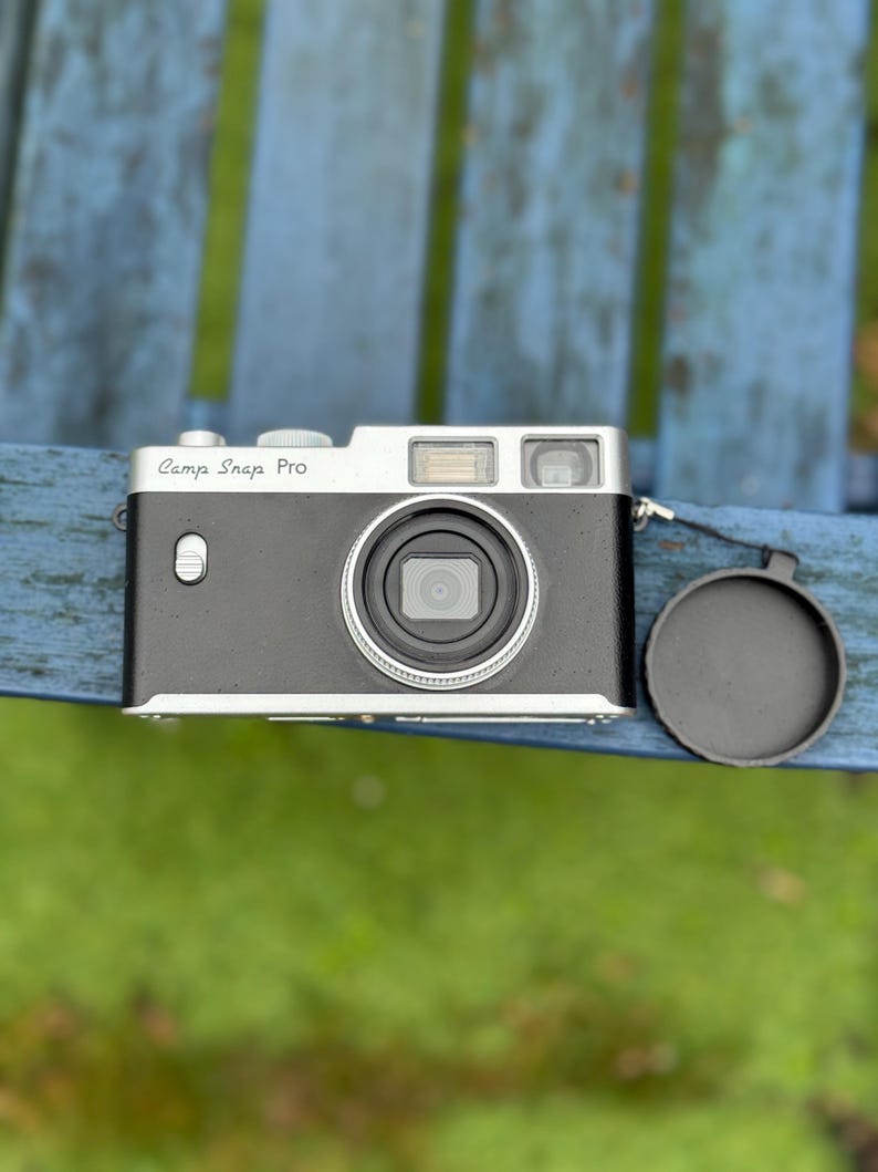 Pode incluir: Uma c&acirc;mera Camp Snap Pro preta e prateada com uma tampa de lente. A c&acirc;mera tem uma parte superior prateada e uma lente proeminente. O texto "Camp Snap Pro" est&aacute; impresso na frente. A c&acirc;mera est&aacute; em uma superf&iacute;cie azul.