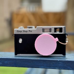 Pode incluir: Uma c&acirc;mera preta e prateada com o texto "Camp Snap Pro" na frente. Uma tampa de lente rosa est&aacute; presa &agrave; c&acirc;mera. A c&acirc;mera est&aacute; em uma superf&iacute;cie azul.