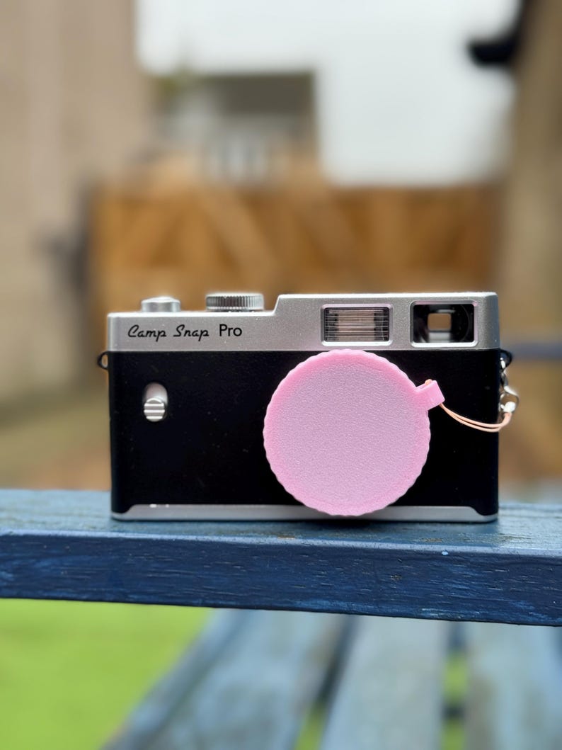 Pode incluir: Uma c&acirc;mera preta e prateada com o texto "Camp Snap Pro" na frente. Uma tampa de lente rosa est&aacute; presa &agrave; c&acirc;mera. A c&acirc;mera est&aacute; em uma superf&iacute;cie azul.