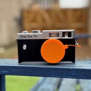 Pode incluir: Uma c&acirc;mara Camp Snap Pro preta e prateada com uma tampa de lente laranja. A c&acirc;mara est&aacute; sobre uma superf&iacute;cie de madeira. A tampa da lente &eacute; redonda e tem uma corda.