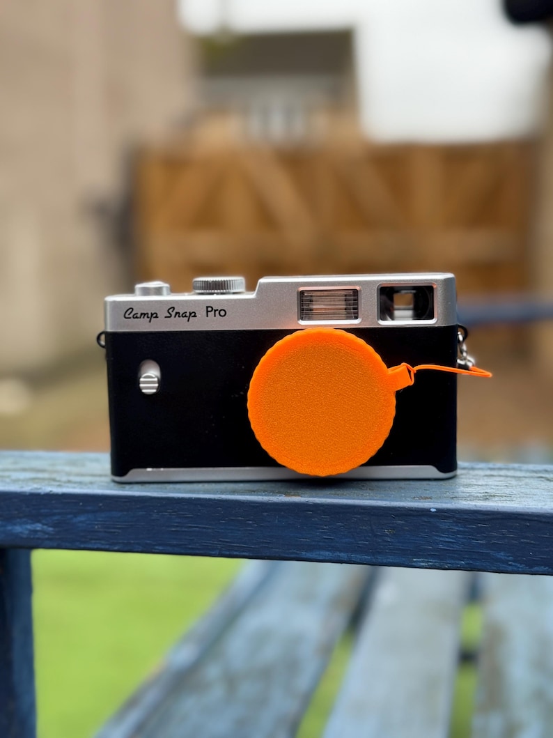 Pode incluir: Uma c&acirc;mara Camp Snap Pro preta e prateada com uma tampa de lente laranja. A c&acirc;mara est&aacute; sobre uma superf&iacute;cie de madeira. A tampa da lente &eacute; redonda e tem uma corda.