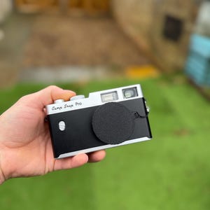 Pode incluir: Uma c&acirc;mera preta e prateada com uma tampa de lente &eacute; segurada em uma m&atilde;o. A c&acirc;mera tem as palavras "Camp Snap Pro" impressas na parte superior. O fundo est&aacute; desfocado, mostrando grama verde e uma porta de madeira.