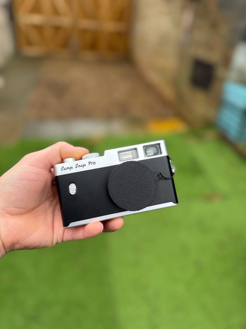 Pode incluir: Uma c&acirc;mera preta e prateada com uma tampa de lente &eacute; segurada em uma m&atilde;o. A c&acirc;mera tem as palavras "Camp Snap Pro" impressas na parte superior. O fundo est&aacute; desfocado, mostrando grama verde e uma porta de madeira.