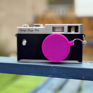 Pode incluir: Uma c&acirc;mera Camp Snap Pro preta e prateada com uma tampa de lente rosa brilhante. A c&acirc;mera &eacute; retangular com uma parte superior prateada e um corpo preto. A tampa da lente &eacute; circular e est&aacute; presa.