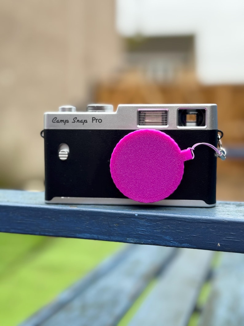 Pode incluir: Uma c&acirc;mera Camp Snap Pro preta e prateada com uma tampa de lente rosa brilhante. A c&acirc;mera &eacute; retangular com uma parte superior prateada e um corpo preto. A tampa da lente &eacute; circular e est&aacute; presa.