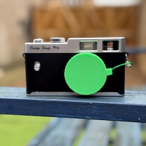 Pode incluir: Uma c&acirc;mera Camp Snap Pro preta e prateada com uma tampa de lente verde brilhante. A c&acirc;mera tem um corpo retangular com uma parte superior prateada e uma frente preta. A tampa da lente &eacute; circular e presa com uma corda verde.