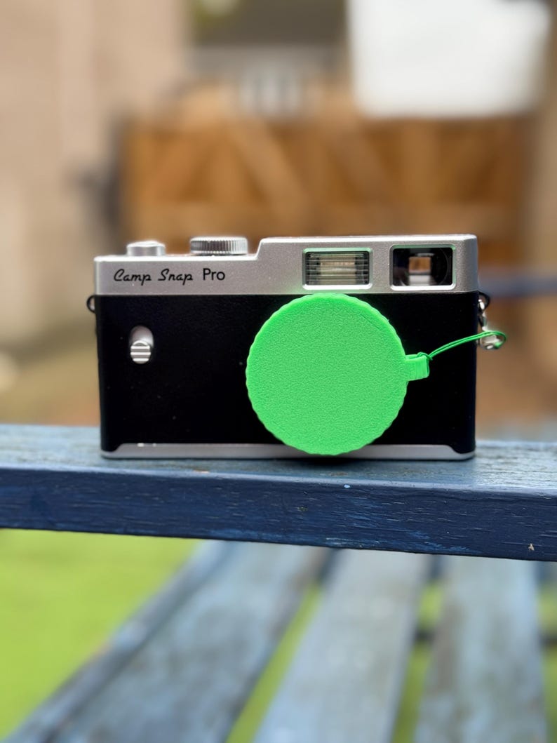 Pode incluir: Uma c&acirc;mera Camp Snap Pro preta e prateada com uma tampa de lente verde brilhante. A c&acirc;mera tem um corpo retangular com uma parte superior prateada e uma frente preta. A tampa da lente &eacute; circular e presa com uma corda verde.