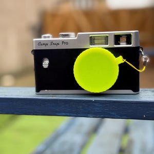 Pode incluir: Uma c&acirc;mera preta e prateada com o texto "Camp Snap Pro" &eacute; mostrada. Uma tampa de lente amarela brilhante est&aacute; presa &agrave; c&acirc;mera. A c&acirc;mera est&aacute; apoiada em uma superf&iacute;cie azul, com um fundo verde desfocado.