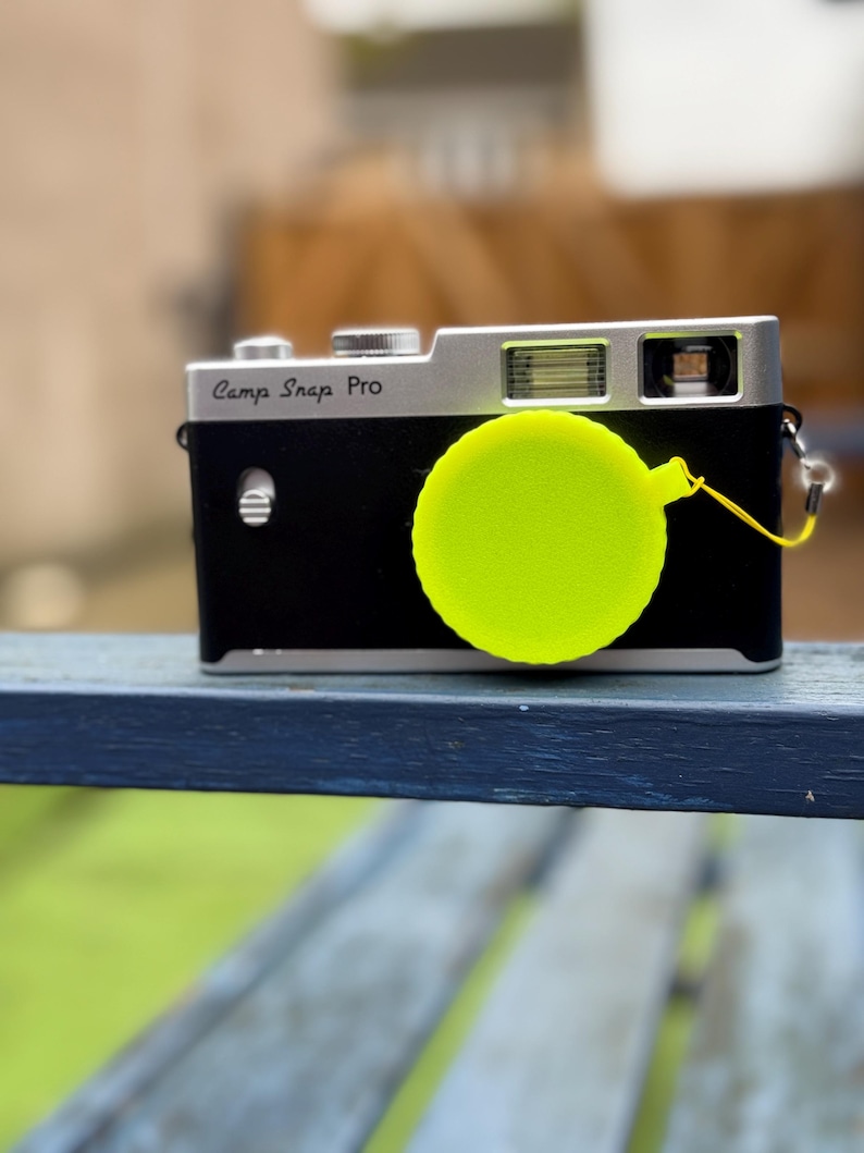 Pode incluir: Uma c&acirc;mera preta e prateada com o texto "Camp Snap Pro" &eacute; mostrada. Uma tampa de lente amarela brilhante est&aacute; presa &agrave; c&acirc;mera. A c&acirc;mera est&aacute; apoiada em uma superf&iacute;cie azul, com um fundo verde desfocado.