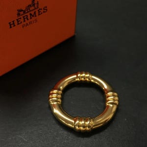 Puede incluir: Un anillo dorado con un diseño circular. El anillo presenta tres juegos de detalles horizontales y acanalados alrededor de su circunferencia. El fondo incluye una porción de una caja naranja con el logotipo "HERMES PARIS".
