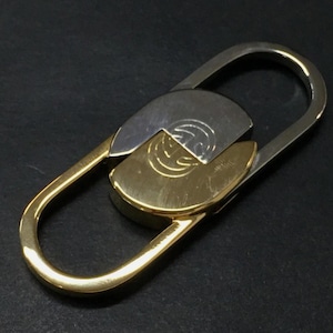May include: A two-tone metal keychain with a gold-colored base and a silver-colored top. The keychain has a unique interlocking design with a circular emblem on the silver portion.