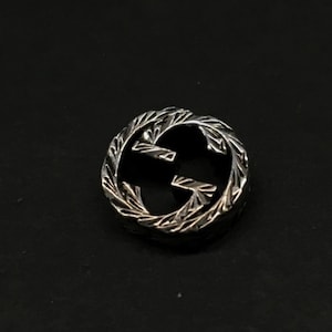 May include: A silver-toned, circular emblem featuring the interlocking 'G' logo. The emblem has a textured, leaf-like border and is set against a dark background. The design is a classic representation of luxury branding.