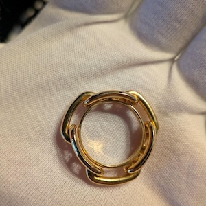 May include: A gold-toned ring featuring a circular band design with three interconnected, smooth, polished metal links that form a continuous, rounded shape. The ring has a reflective, metallic finish and a simple, uniform structure.