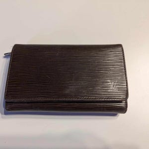 May include: A brown leather wallet with a textured, ridged surface. The wallet has a flap closure and a small zipper. A logo is visible on the front. The wallet is rectangular and appears to be made of high-quality leather.