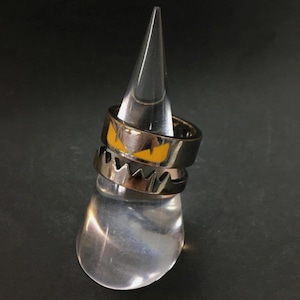 May include: Two rings with a Halloween theme are stacked on a clear display stand. The top ring is black with yellow eye-shaped designs. The bottom ring is silver with a jagged edge.