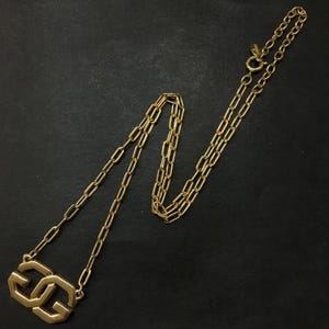 Puede incluir: Collar dorado con un colgante con doble G. El collar presenta una delicada cadena y un colgante geométrico con G entrelazadas. La joya se exhibe sobre un fondo oscuro, resaltando su brillo metálico.