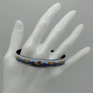 Puede incluir: Una pulsera decorativa de esmalte con un diseño repetido de leones, árboles y montañas. La pulsera tiene un interior negro y un exterior blanco con detalles en azul y amarillo. La pulsera se muestra en una mano blanca.