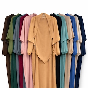 Könnte beinhalten: Eine Kollektion fließender, einfarbiger Kleider in verschiedenen Farbtönen, darunter Braun, Grün, Rosa und Blau. Jedes Kleid hat ein passendes, schalartiges Oberteil, wodurch ein koordiniertes Ensemble entsteht. Die Kleidungsstücke werden auf Kleiderbügeln vor weißem Hintergrund präsentiert.