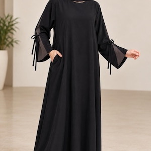 May include: A floor-length black abaya dress featuring long sleeves with split cuffs and tie-string details. The garment includes side pockets and is paired with a matching black headscarf. The design is simple, with a loose, flowing silhouette.