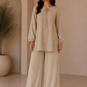 May include: A beige two-piece outfit. The long-sleeved top has a tie-neck detail and the wide-leg trousers have a flowing silhouette. The top features a subtle floral pattern. The outfit is made of a soft, flowing fabric.