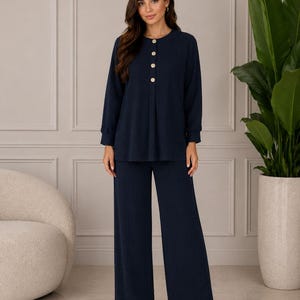 May include: A dark navy blue two-piece set consisting of a long-sleeved top and wide-leg trousers. The top features a round neckline with four light-colored buttons down the center front and cuffed sleeves. The fabric has a subtle vertical texture.