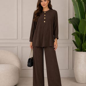 May include: A dark brown two-piece set consisting of a long-sleeved tunic top with four light-coloured buttons along the neckline and matching wide-leg trousers. The fabric features a subtle vertical ribbed texture throughout the entire garment.