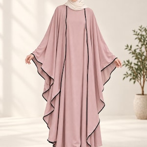 Può includere: Un abaya lungo fino ai piedi color rosa polvere, composto da un vestito interno senza maniche e uno strato esterno fluido a mantella con profili neri lungo i bordi. L'abito è abbinato a un hijab color crema e scarpe chiare a punta.