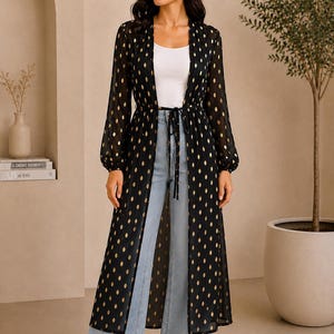 May include: Black duster coat with gold dot pattern, long sleeves, and a tie waist. The coat is worn over a white top and light blue wide-leg jeans. A stylish, long, open-front garment.