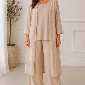 May include: A three-piece beige ensemble consisting of a sleeveless top, wide-leg trousers, and a long, open-front duster cardigan. The duster features a subtle, tonal floral pattern throughout, while the top and trousers are solid beige.