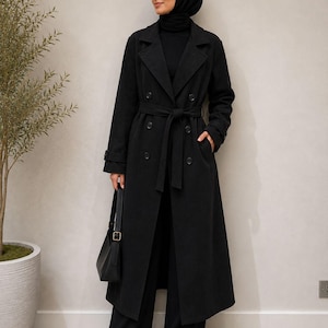 Könnte beinhalten: Ein langer, schwarzer, zweireihiger Trenchcoat mit fallendem Revers, einem passenden Bindegürtel in der Taille und langen Ärmeln mit geknöpften Riegeln an den Manschetten. Der Mantel wird über einer schwarzen Hose getragen und mit einer schwarzen Tasche kombiniert.