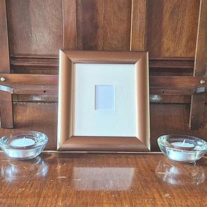 May include: A brown wooden picture frame with a white mat and a small square opening. Two clear glass votive candle holders with lit candles sit on a wooden surface. The background is a dark wooden cabinet.