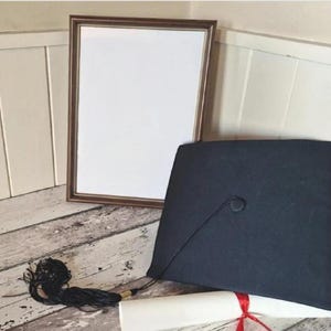 May include: A black graduation cap with a tassel, a rolled diploma, and a framed blank paper are displayed. The cap has a button on top. The diploma is tied with a red ribbon, and the frame is brown.