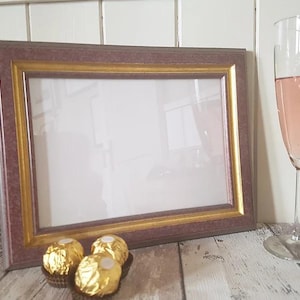 May include: A brown picture frame with a gold inner border. The frame is sitting on a wooden surface with three gold-wrapped chocolates in front of it. A glass of pink wine is in the background.