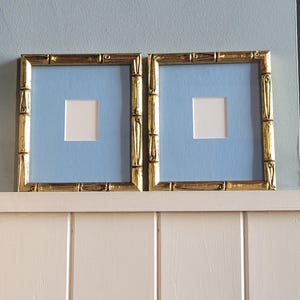 Peut inclure: Deux cadres carrés avec des cadres dorés de style bambou. Chaque cadre a un passe-partout bleu clair avec une ouverture carrée blanche. Les cadres sont exposés sur une étagère blanche contre un mur bleu clair.