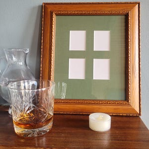May include: A wooden picture frame with a green mat and four empty openings. The frame has a decorative border and is sitting on a wooden surface. A glass of amber liquid and a small white candle are also on the surface.