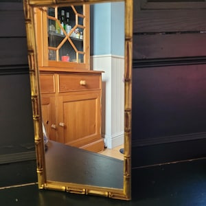 Peut inclure: Un miroir encadré d'or avec un motif de style bambou. Le miroir reflète une armoire en bois avec des portes et des tiroirs en verre.
